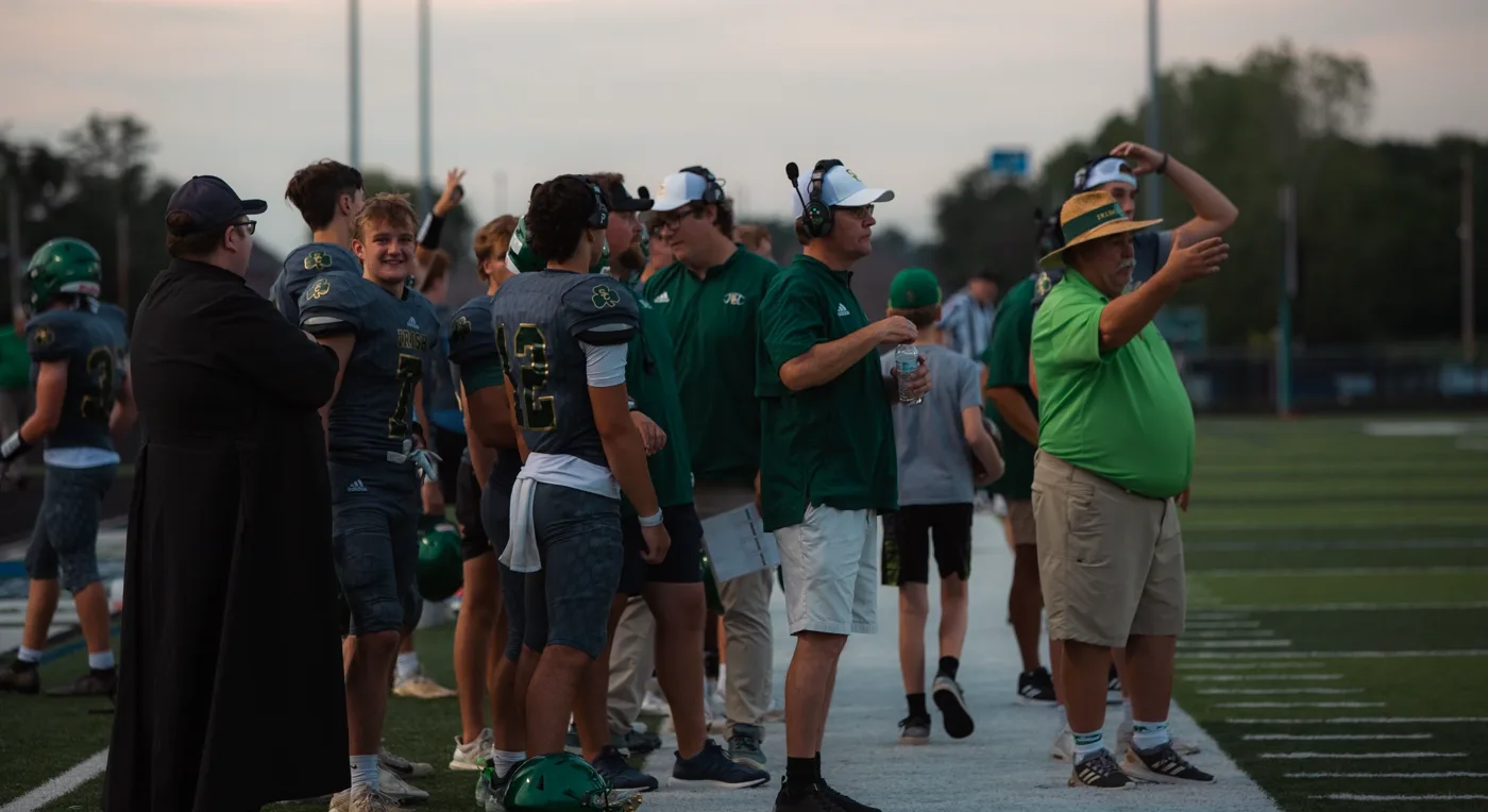 Coaches and players gathered on a high school football sideline during an evening game